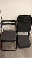 Four black folding chairs arranged with one chair open and others folded. Chairs have metal frames, plastic backs, and grey cushions.