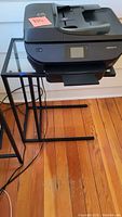 Side table with a black metal base and clear glass top, with printer partly on top for scale, hardwood floor background.
