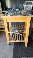 Front view of the wood butcher block cart with two slatted shelves. Top shows some visible scratches and a wooden crack. Two vintage cookbooks placed on the bottom shelf. Assorted metal hardware seen in background but not included.