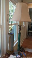 Two vintage style lamps side by side near window, one with beige fabric shade and greenish-bronze column base, other with silver cylindrical base and no shade.