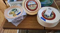 Set of Mottahedeh fruit decorated plates stacked beside Le Troupeau plates in box on wooden table