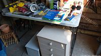 Folding table with various items on the surface and small white drawer unit underneath. Table surface white plastic with metal legs visible underneath.
