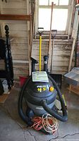 Shop Vac QULP550 14.5 gallon wet/dry vacuum with attached hose, leaf rakes, snow shovels, rough scrub brush, and extension cords arranged in garage
