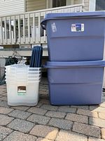 Two large stacked blue 45 gallon rubbermaid Roughneck totes with lids and five smaller clear plastic totes with their lids, arranged on a stone paved surface near a house porch.