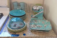 Photo of vintage style juicer, turquoise 222 Fifth fine china plates and mugs, clear tall glasses, food cover dome, and multiple patterned plates including Villeroy and Boch and other brands on kitchen counter.