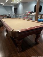 Large wooden pool table with tan felt, showing corner and side pocket and carved legs.