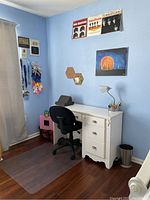 Full view of white girls desk with three drawers and black office swivel chair in a bedroom with light blue walls and hardwood floor.