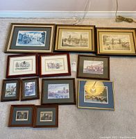 Twelve framed prints arranged on floor showing variety of cityscape and architectural drawings in various frame styles and sizes.