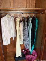 A closet rod holding seven women's garments including jackets, blouses, and cardigans in various colors such as white, yellow, neutral, grey, aqua, and dark green. A handbag sits below on the floor.