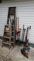 Photo showing two wooden step ladders side by side against a white siding wall with several garden tools nearby, including shovels and a Black and Decker electric edger.