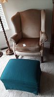 Beige highback wing chair with blue ottoman in front. Chair fabric appears velvety.