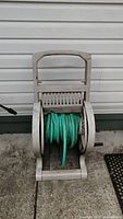 Front view of green garden hose coiled on beige Suncoast plastic reel cart against white house siding on concrete.