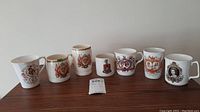 Seven white commemorative china mugs and cups arranged side by side on a wooden surface, showing variety of British royal crests and designs.