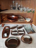 Wide view of two wooden shelves and floor displaying a collection of glassware on the upper shelf, wooden and stainless steel serving ware on the lower shelf and on the floor in front, including a large wooden bowl, trays and divided plates.
