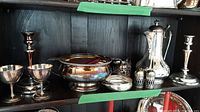 Wide view of shelf holding entire silverplate collection showing coffee pot, candle holders, goblets, bowls, sugar bowl and salt & pepper shakers. Items have tarnish and surface wear.