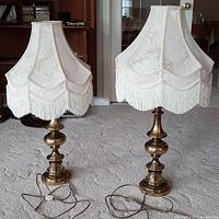 Pair of brass table lamps with embroidered cream fabric shades and white fringe, standing on carpeted floor.