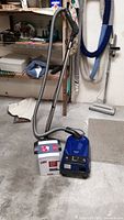 Blue Miele S251i vacuum cleaner with hose and metal wand upright on concrete floor, with box of bags beside it.