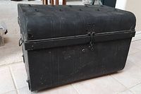 Side angled view of black metal trunk showing rounded top, latch, and wear marks.