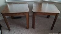 Pair of wooden end tables side by side on carpeted floor under window with natural light, showing general condition and matching design