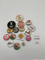 Photo showing a full view of the 16 assorted vintage pinback buttons with various designs including Playboy bunny, Lobster, OK text, and others.