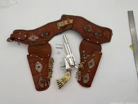 Full view showing dark brown rawhide holster and matching belt decorated with silver diamond-shaped conchos with red bead centers, and silver toy cap gun with striped handle overlaying the holster.