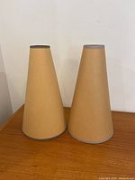 Two tall, conical brown paper lamp shades with gray top and bottom trim banding, standing upright on a wood surface against a white wall.