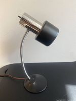 Photo of the lamp showing the chrome cylindrical head with black shade and flexible gooseneck attached to a round black base.