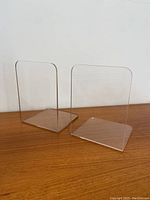 Pair of clear acrylic bookends shown from front angle on wooden surface against white wall.