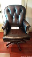 Front view of dark brown leather office chair showing button tufted backrest and seat, rivet details on armrests, wooden base with casters.