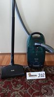 Front view of green Miele Monte Verde Electro Plus canister vacuum with attached hose and floor head on dark patterned carpet.