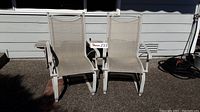 Front view of two matching beige metal frame patio rocking chairs with fabric seats and attached wooden side trays.