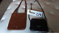 Two shoulder bags side by side on a mattress: brown Radley London leather bag with long adjustable strap, black Brighton bag with long thin strap and front flap pocket with logo clasp.