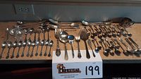 Wide view of all flatware pieces laid out on counter showing two rows of spoons, forks, serving pieces, and souvenir spoons including small and large utensils.