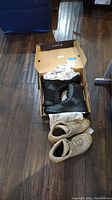 Photo showing two pairs of boots side by side on wooden floor, tan Lowa boots without box and black Law Pro boots inside original box with white tissue paper.