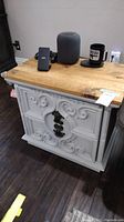 White wood end table with plain natural wood top, small decorative metal handle, and ornate carved design on front. Scratches visible on wood surfaces.