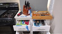 View of four white kitchen drawers pulled open revealing flatware including spoons, forks, knives and plastic cooking utensils such as spatulas and serving spoons.