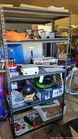 Photo showing tall shelving unit with mixed hardware items, small containers, boxes, and household supplies including fishing reel and thermostat plate.