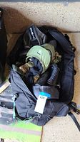 Photo showing black duffel bag containing green tactical paintball helmet, camouflage fabric, and air canister.