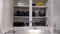 Kitchen cabinet with white large bowl on top shelf, blue mugs in the middle, and dark blue plates at bottom shelf