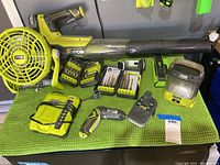 Overhead view of Ryobi cordless blower, fan, drill, batteries, charger, bit sets and work light arranged on green mat