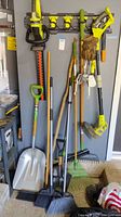 Wide view of the yard tools hanging on a yellow rack and resting on the floor, showing trimmer, shovels, rakes, brooms, and gloves.