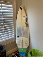 Full view of the surfboard leaning against a wall near a window showing the entire board and its condition.