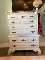 Front view of tallboy dresser showing multiple drawers with brass handles and white paint finish, with a TV and decorative items on top.