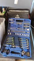 Full view of blue plastic Husky tool case open showing combination wrenches, bits, sockets, ratchet handle, and screwdriver inside. Husky tool bag in background.