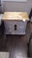 Front view of white wood end table showing natural wood plank top and carved front panel with dark decorative metal accent.