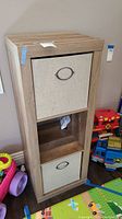 Front view of wood storage unit with 2 beige fabric bins and open middle shelf against wall, surrounded by children's toys.