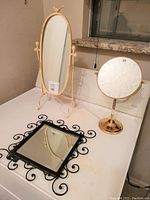 Photo showing three table top personal mirrors: oblong cream frame mirror on stand, black square mirror with wrought iron frame, and round gold magnifying mirror.