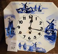 Front view of the porcelain clock face featuring blue and white Delft-style paintings of windmills, boats, and houses. Visible crack on the clock's lower front.