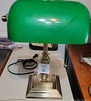 Front view of the banker's lamp showing green glass shade, brass base, tag with lot number, power cord.
