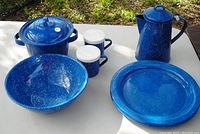 Blue speckled enamel ware dish set displayed outdoors on white table with grass background. Includes plates, coffee pot, bean pot with lid, large bowl, and salt & pepper shakers.